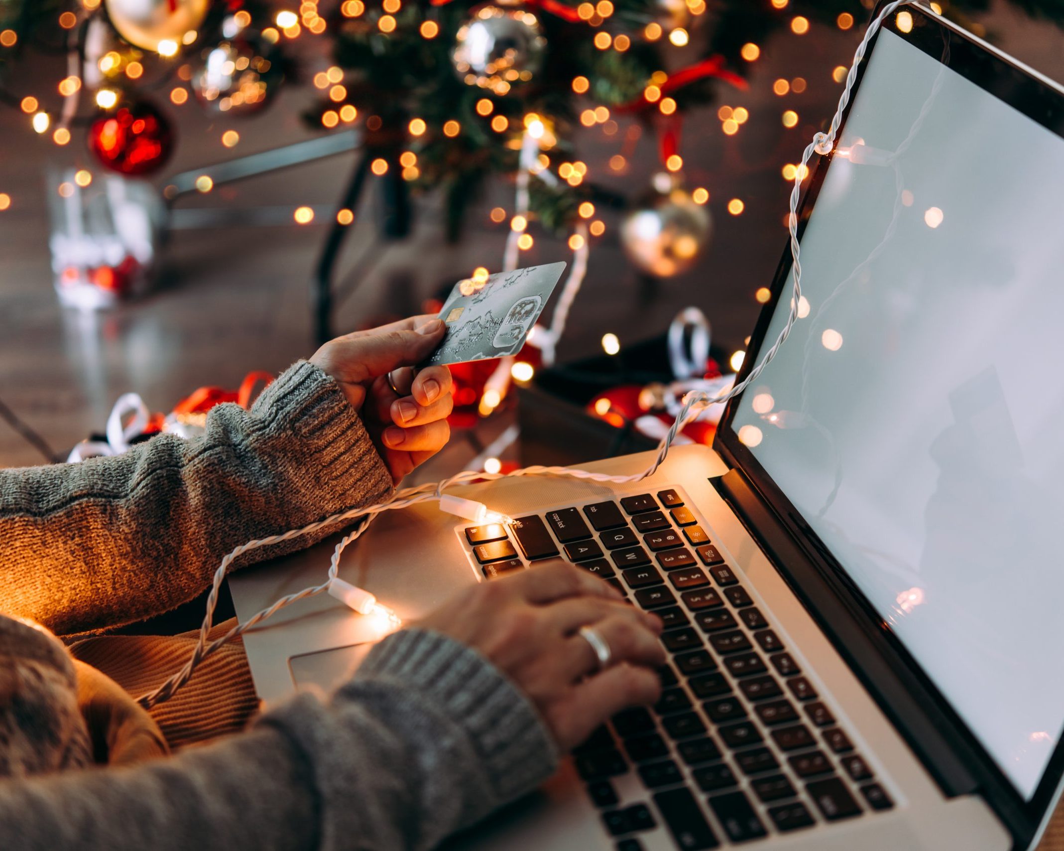 female hand typing on a laptop while holding a credit card with christmas lights in the background