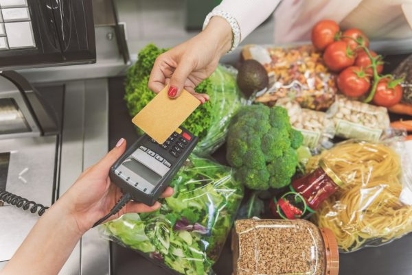 Customer is standing near cash register and giving bank card to cashier. Close up of female hand holding means for contactless payment. Different goods on tape. Top view
