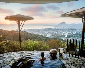 a person sits in an infinity pool overlooking mountains and an ocean beyond
