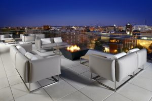 Sofas sit on a hotel rooftop overlooking a city, providing an example of what Cambria hotels are like