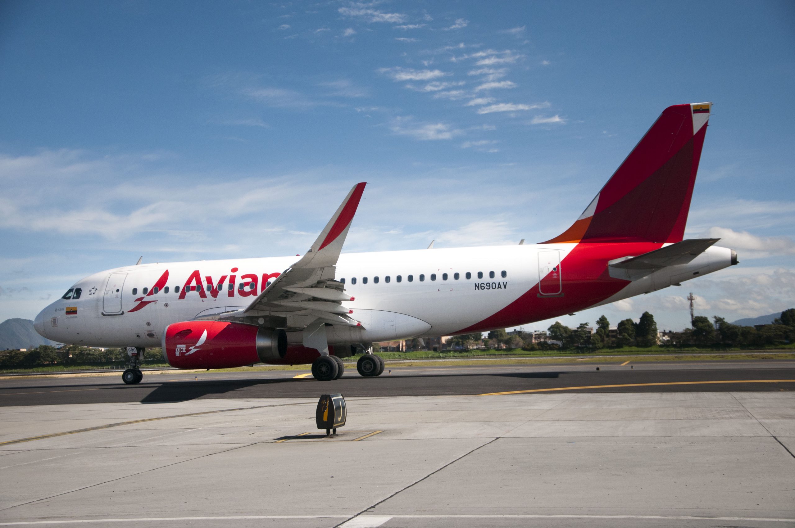 A red-and-white Avianca Airbus A319 sits at an airport with a cloudy, blue sky as a backdrop. (Image courtesy of Avianca)
