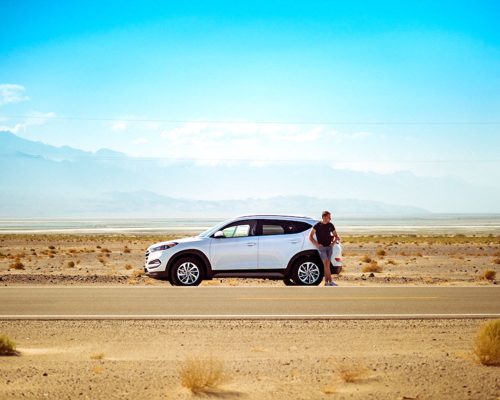 a man stands next a car at the side of a desert highway
