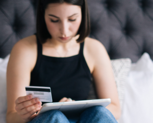 a student sits on her bed holding a credit card and looking at a tablet