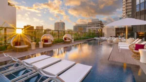 four white lounge chairs in the foreground with a pool in the middle ground and buildings in the background