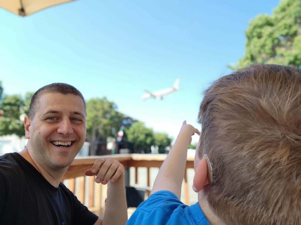 a father takes a selfie of himself and his soon who is pointing to a plane in the sky