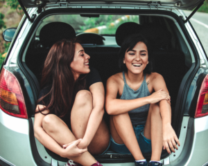 2 friends laugh while sitting in the hatch of a car during a pause on their road trip