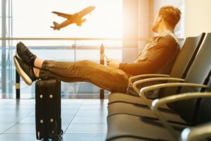 man with feet resting on a suitcase staring out an airport window with a plane taking off at sunset