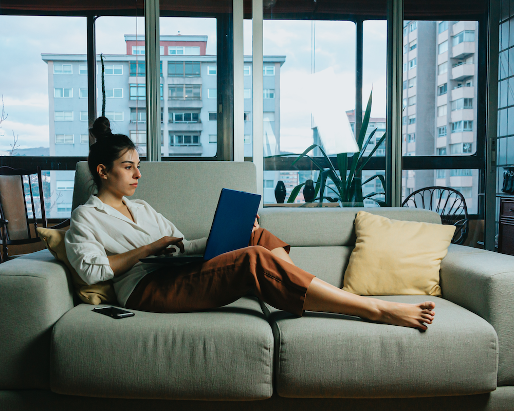 a woman sits on the couch in her apartment, using a laptop