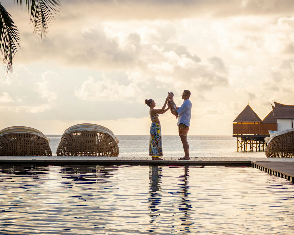 2 parents stand next to a resort hotel pool holding a baby at sunset