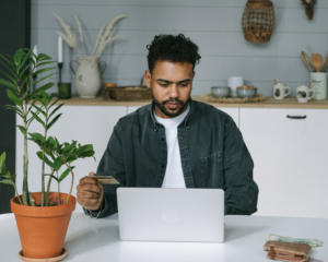 a man sits at a table holding a credit card and looking at his computer
