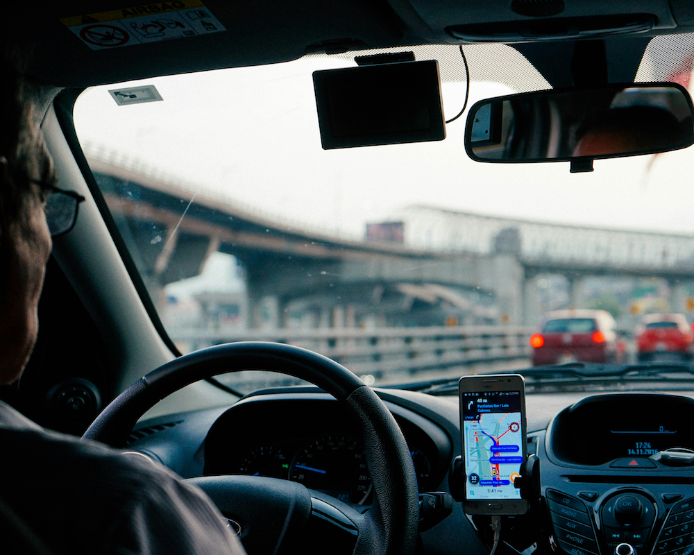looking from the back seat through the front of a car, showing traffic ahead and a driver using a rideshare app