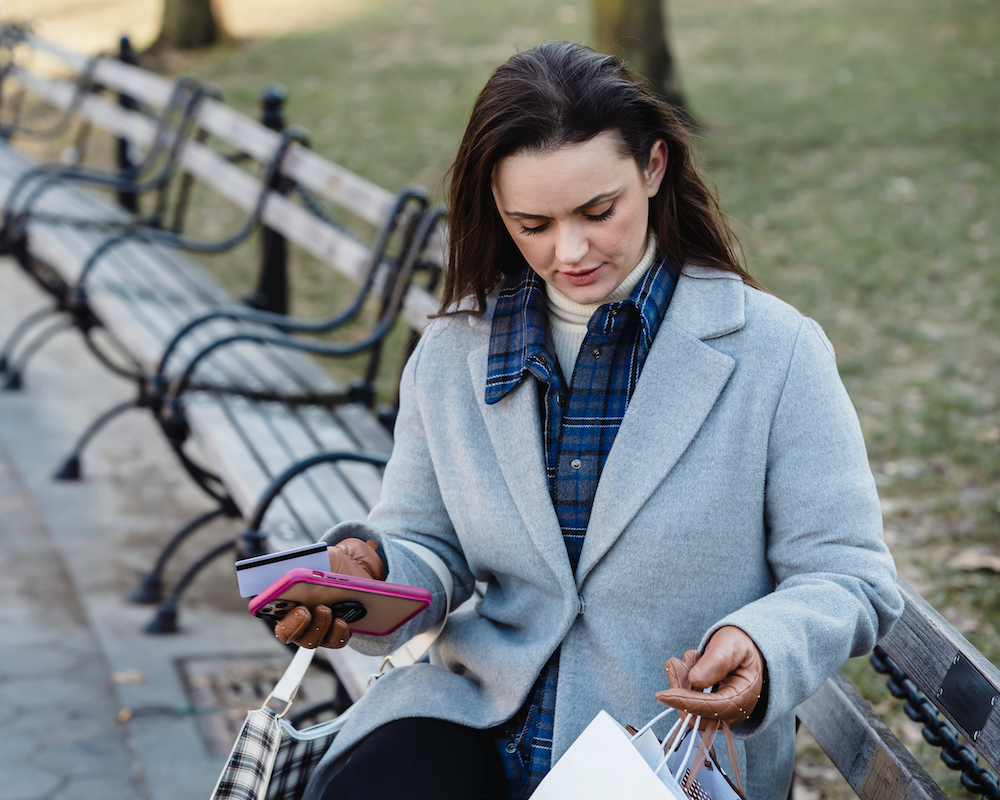 a woman sitting on a park bench holding a smart phone, credit card, and shopping bags