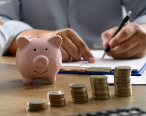A person in the background tracking their finances with a piggy bank and coins in the foreground.