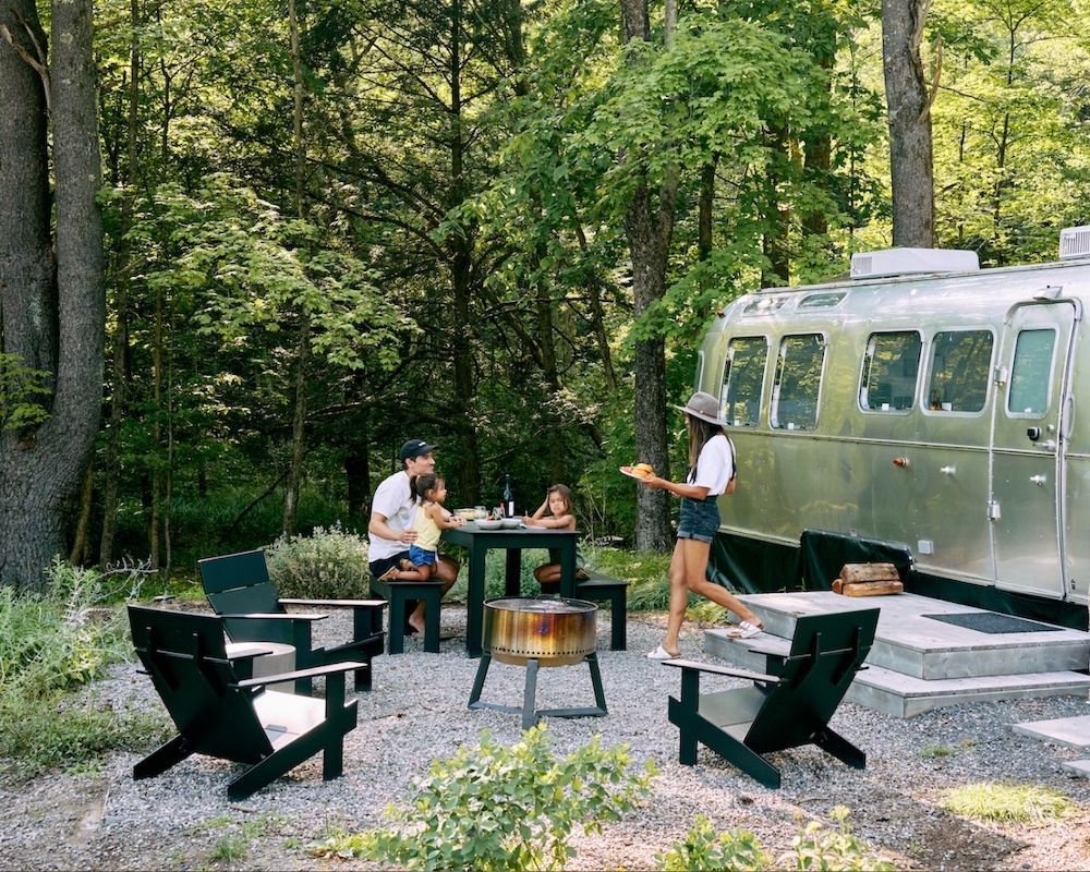 A family at a table and chairs next to a silver Airstream RV at a camp site
