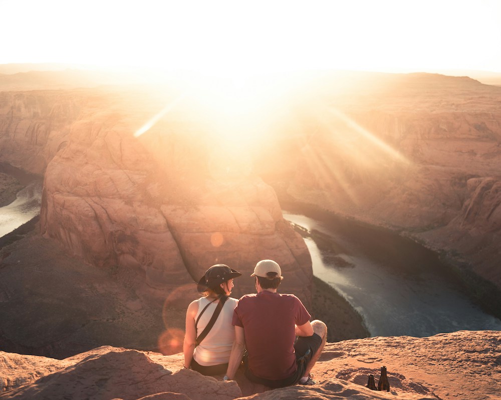 a couple sits on a cliff overlooking a river and a canyon at sunset