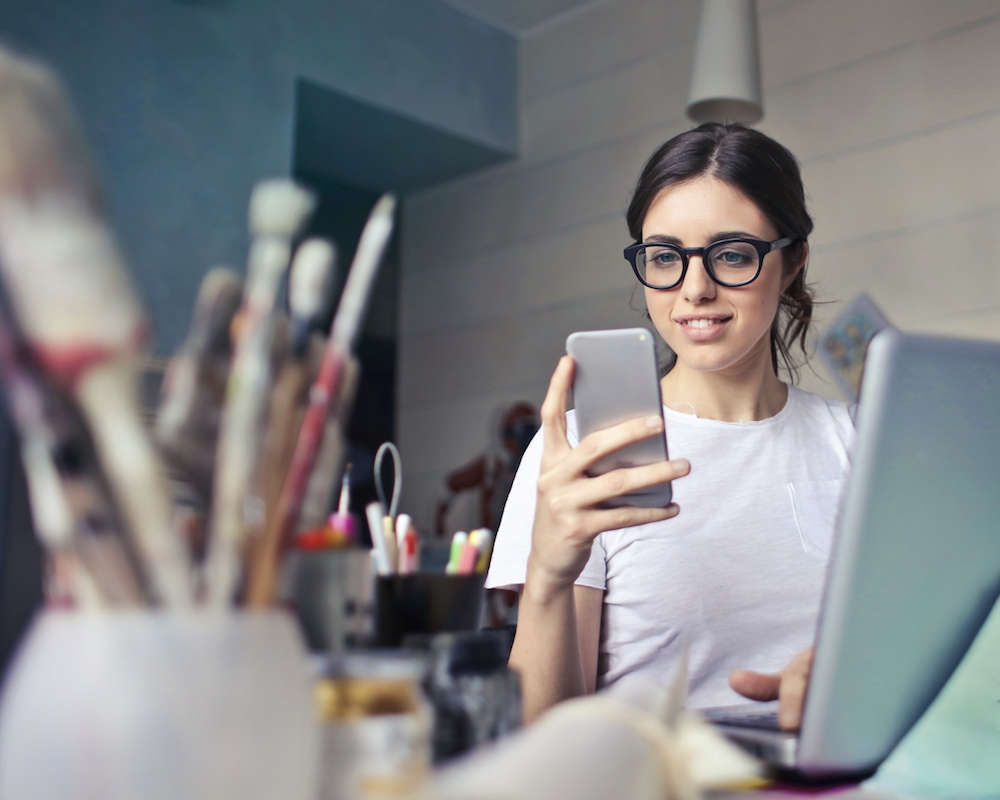 Woman looking at a phone while sitting in front of a laptop.