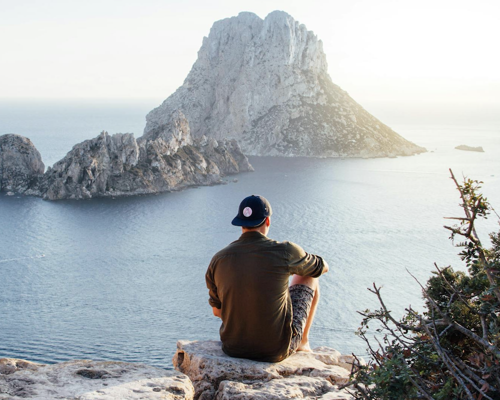 Person sitting on a rock overhang in front of the ocean.