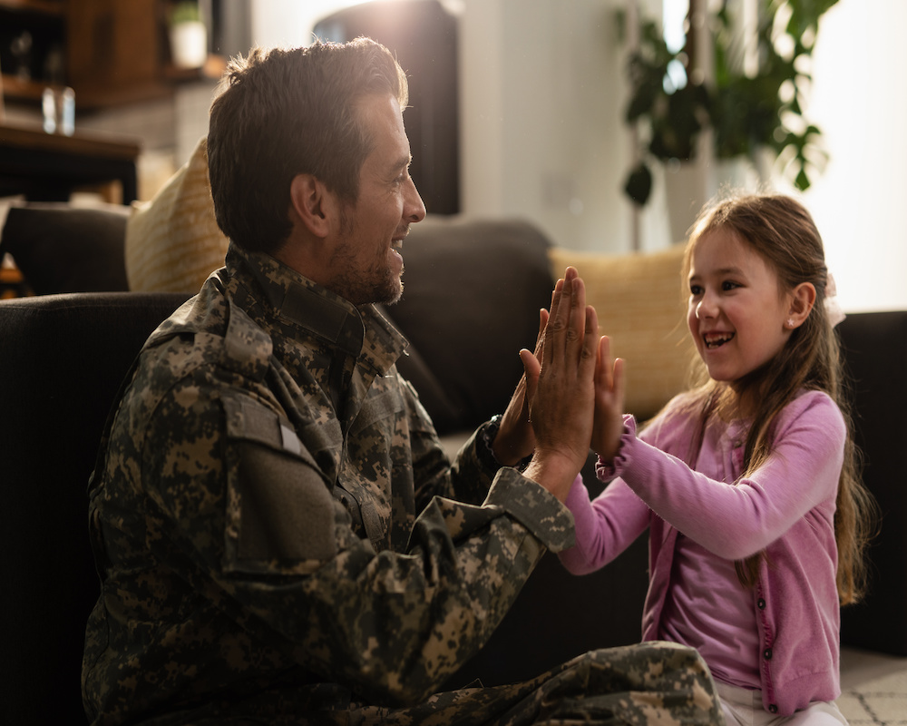 Happy little girl having fun with her military father while playing clapping hands game at home.