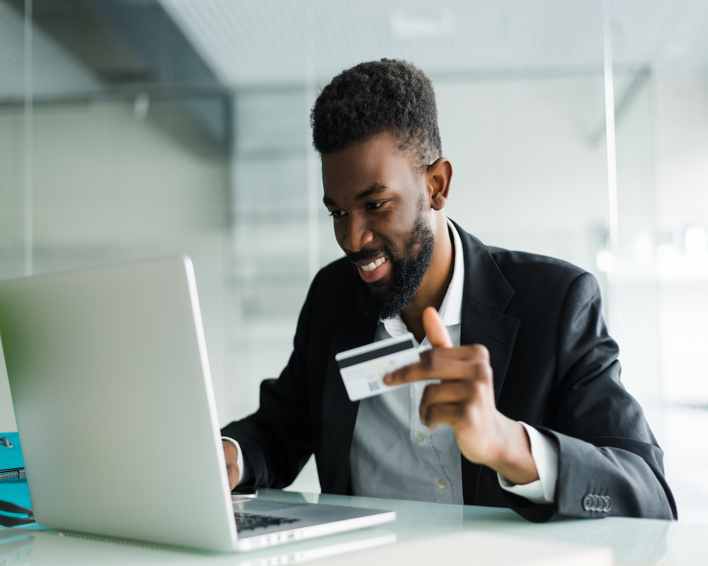 A young man holds a credit card in his left hand while smiling and looking at a laptop screen.