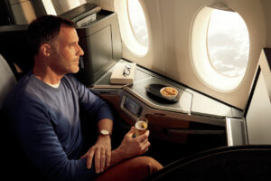 A passenger sitting in a British Airways Club World Suite looks out the window of an aircraft while holding a glass of champagne.