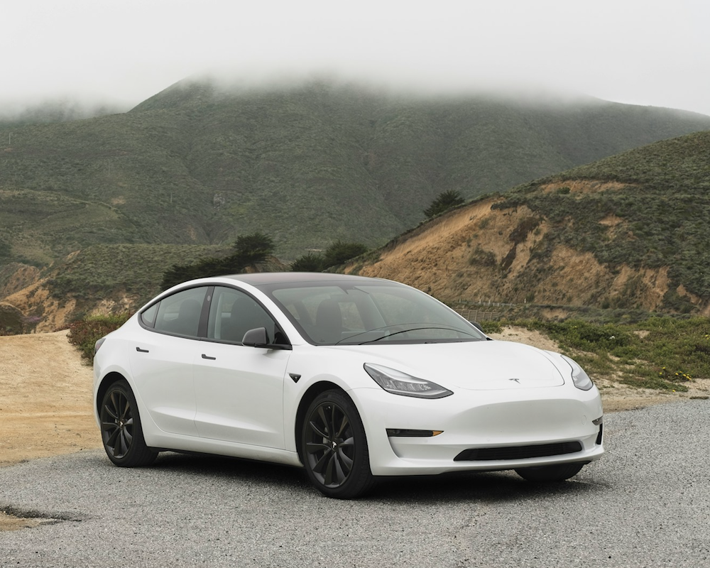 White Tesla Model 3 parked in front of a grassy hill.