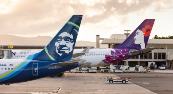A photo shows two jets at gates, one Alaskan and one Hawaiian airlines
