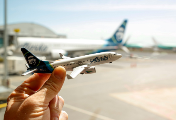 A hand holding a miniature Alaska Airlines plane in front of a full-size Alaska Airlines plane.