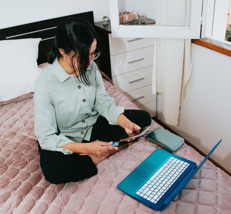 A woman is sitting on her bed with a laptop, credit card and her phone