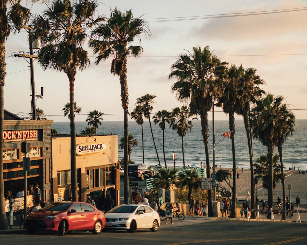 Vehicles parked near a tree and building with the sun setting over Manhattan Beach, California in the background.