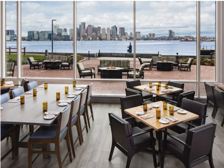 Dining tables inside and outside of glass windows overlooking a harbor with city skyline
