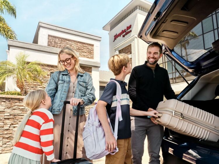 A man and woman help a young boy and girl with getting their luggage out of the truck on a car in front of a Hampton Inn