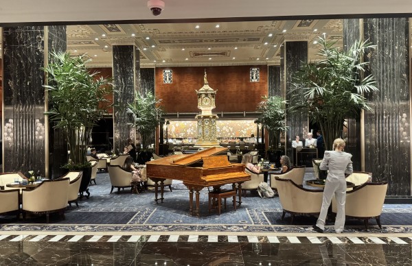 The grand lobby area of the Waldorf Astoria. There is a grand piano, a gold large standing clock and lounge chairs. You can also see a host stand with two employees standing. 
