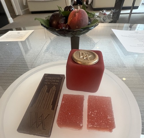 A plate with a chocolate decorated with a imge of the front of the Waldorf astoria, pink fruit jellies, a square red velvet chocolate bite, and a small bowl of fruit.