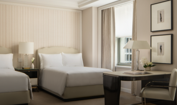 Two double beds in a luxurious but simple room decorated in white and cream colors. There is a desk in the foreground and a window with open cream curtains.
