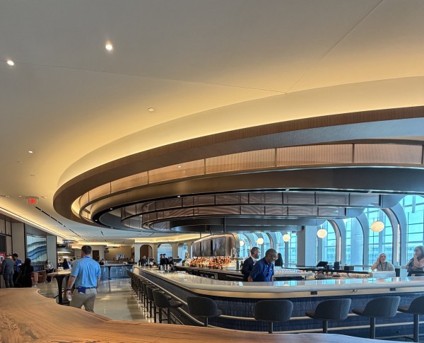 Guests and staff gather around the expansive bar at the new United Club at Houston IAH, featuring sweeping curved ceiling details, modern seating, and floor-to-ceiling windows .